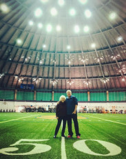 Barbara and Dennis Giacino standing on the field in Manley Field House. Today, it's a practice facility for the Syracuse Orange football team, but when Barbara attended, it was the site of men's basketball games.